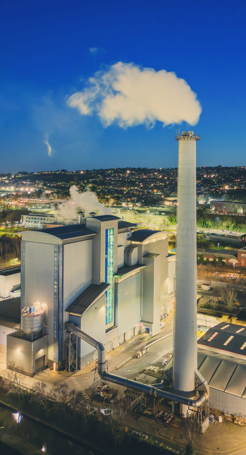 An industrial plant at dusk with a tall stack emitting a white plume above a lit cityscape