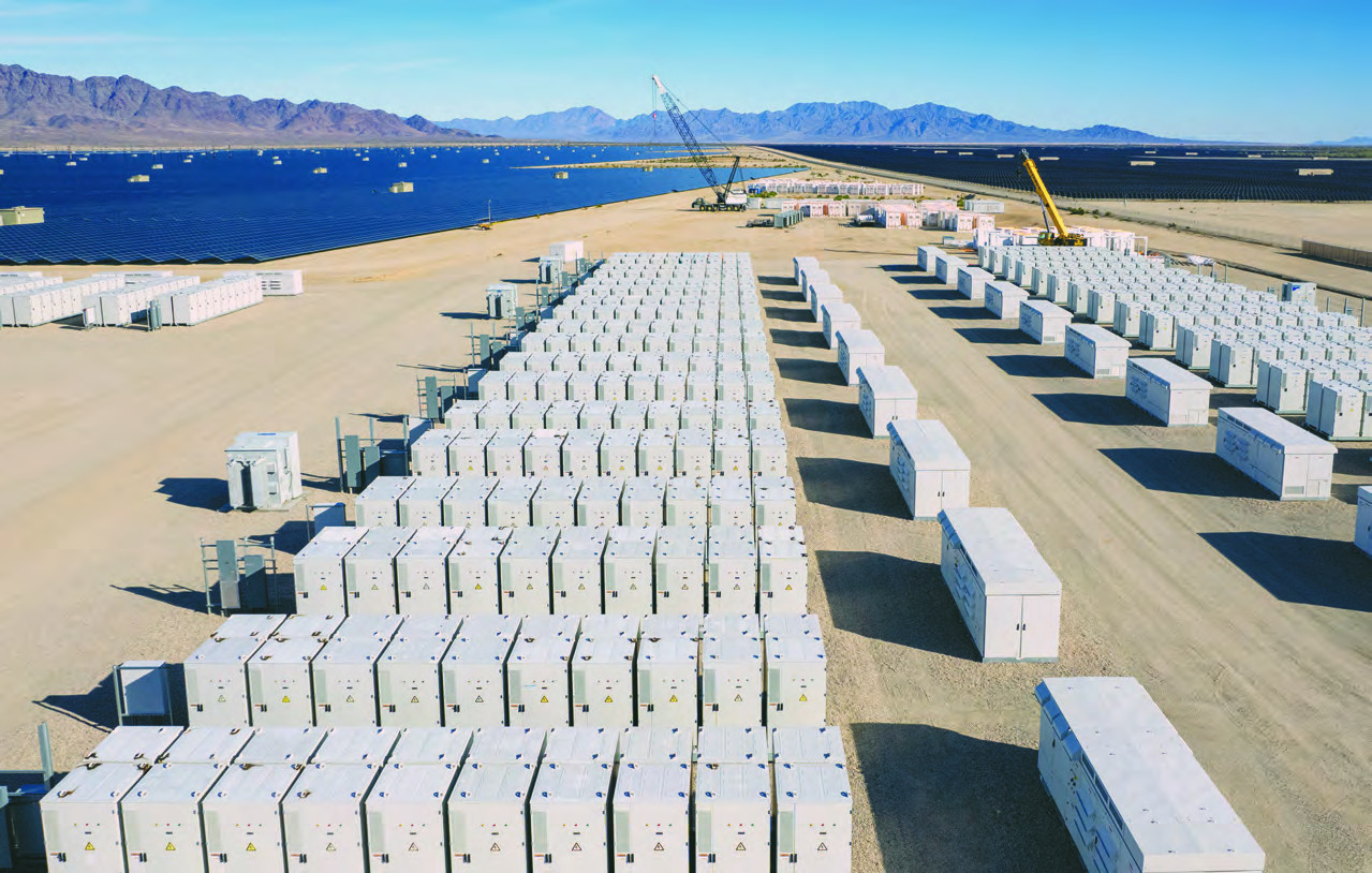 Aerial view of rows of white battery storage containers under construction beside a large desert solar array