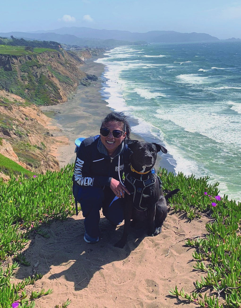 Connie Chern crouching beside a black dog on a coastal cliff path above the ocean
